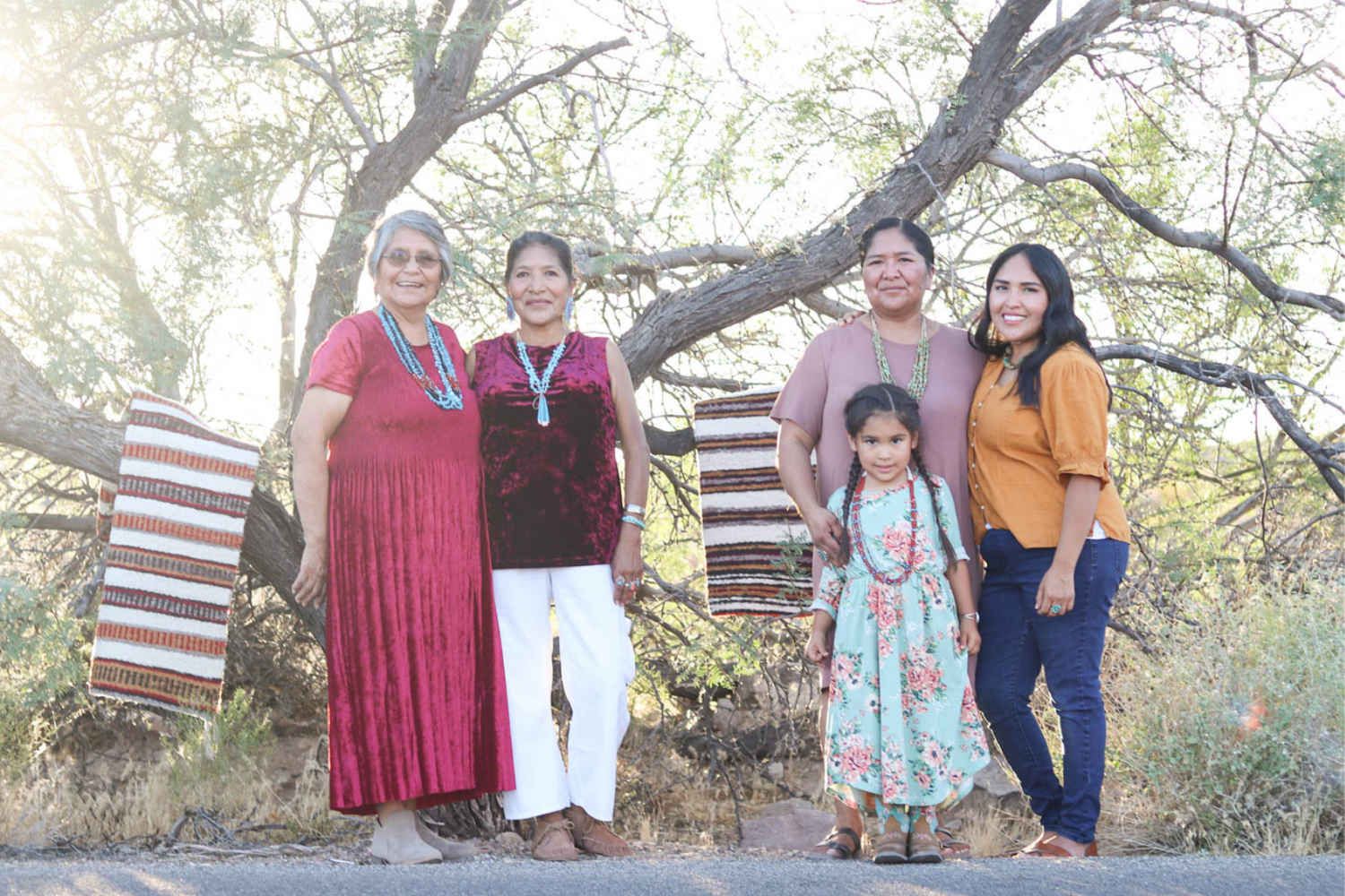 A group of Navajo woman stand and smile in the woods.  